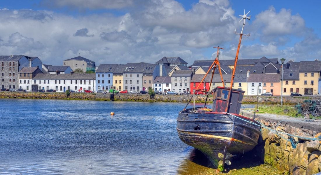 A small rowing boat in the foreground with Galway city in the background with a long row of colourful houses.