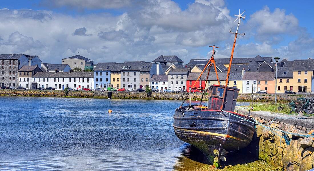 A small rowing boat in the foreground with Galway city in the background with a long row of colourful houses.