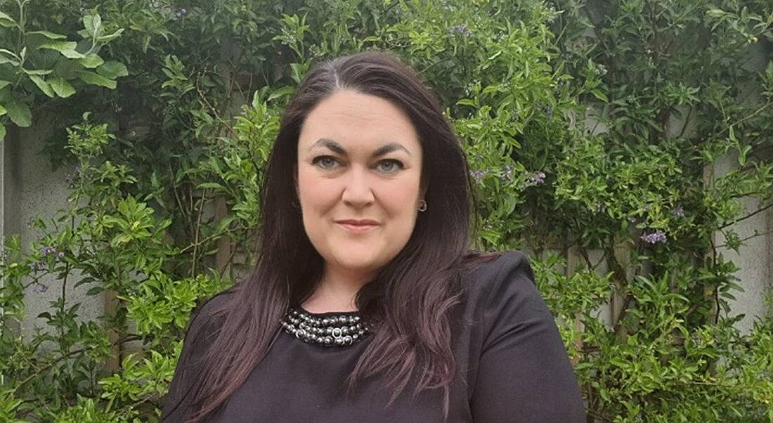 A woman with long brown hair smiles at the camera with greenery in the background. She is Grace Hayes of Mastercard.