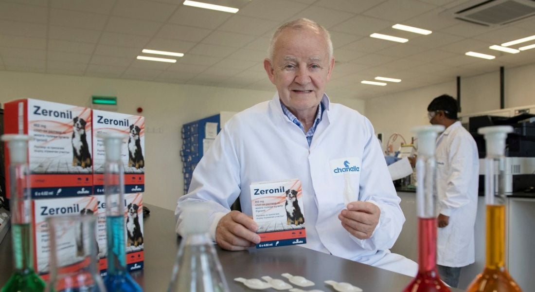 Michael Burke, founder of Chanelle Pharma, is in a lab holding Chanelle products. He is wearing a white lab coat and smiling into the camera.