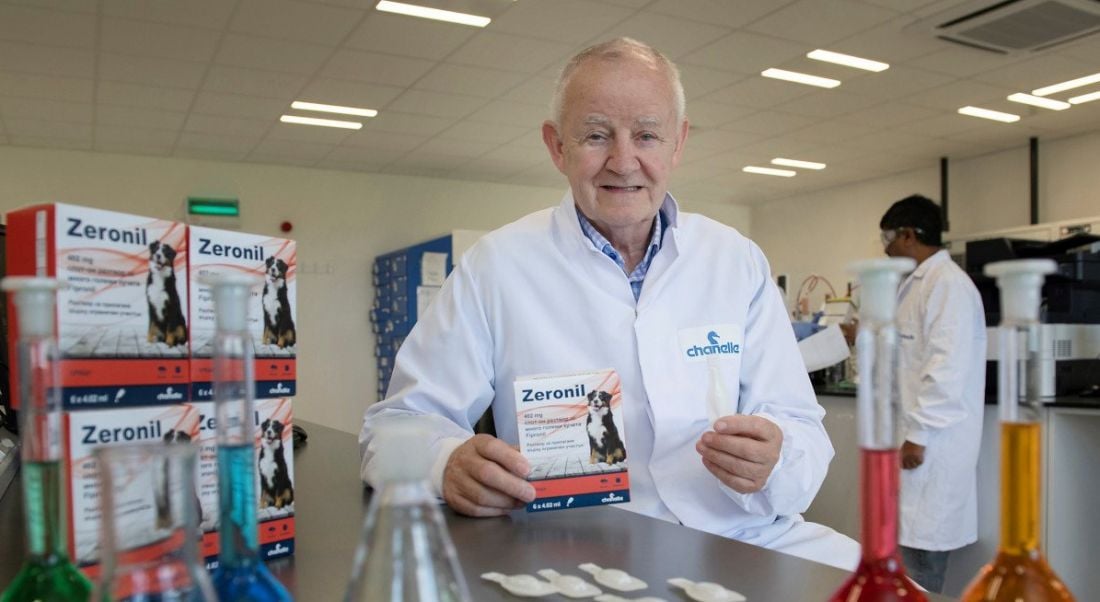 Michael Burke, founder of Chanelle Pharma, is in a lab holding Chanelle products. He is wearing a white lab coat and smiling into the camera.