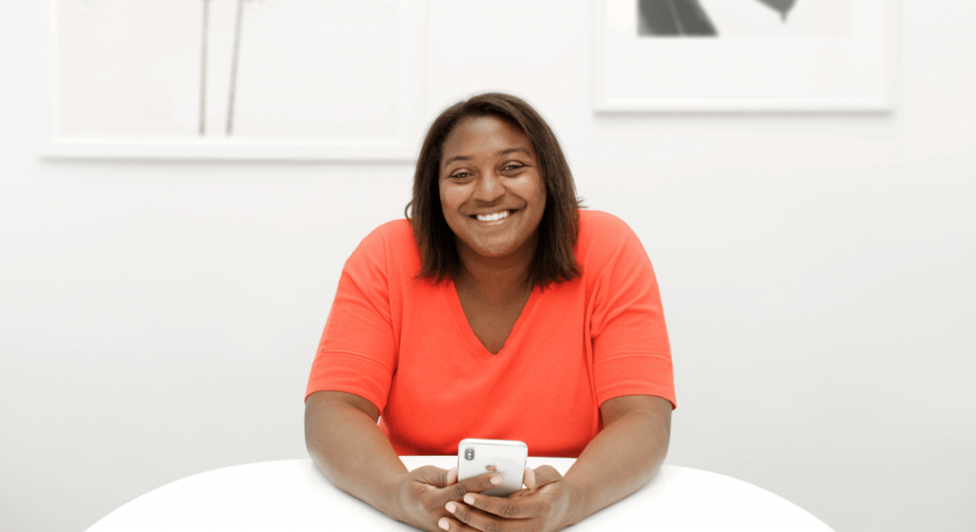Emuye Reynolds of Superhuman is sitting at a white desk in a white-painted room, holding a smartphone while she smiles into the camera. She is wearing a peach-coloured top.