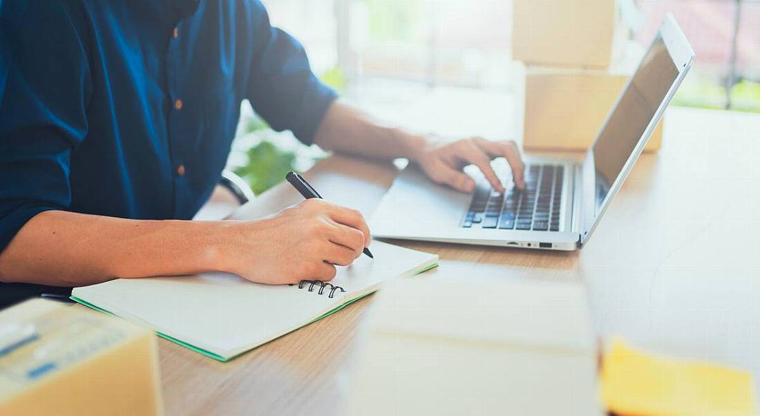 A person working on a laptop at a desk while taking notes in a notebook in a brightly lit office.