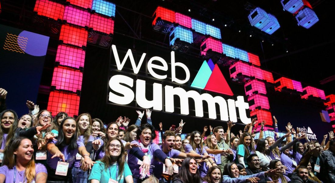 A crowd of people standing together and cheering on a stage decorated with the Web Summit logo and branding.