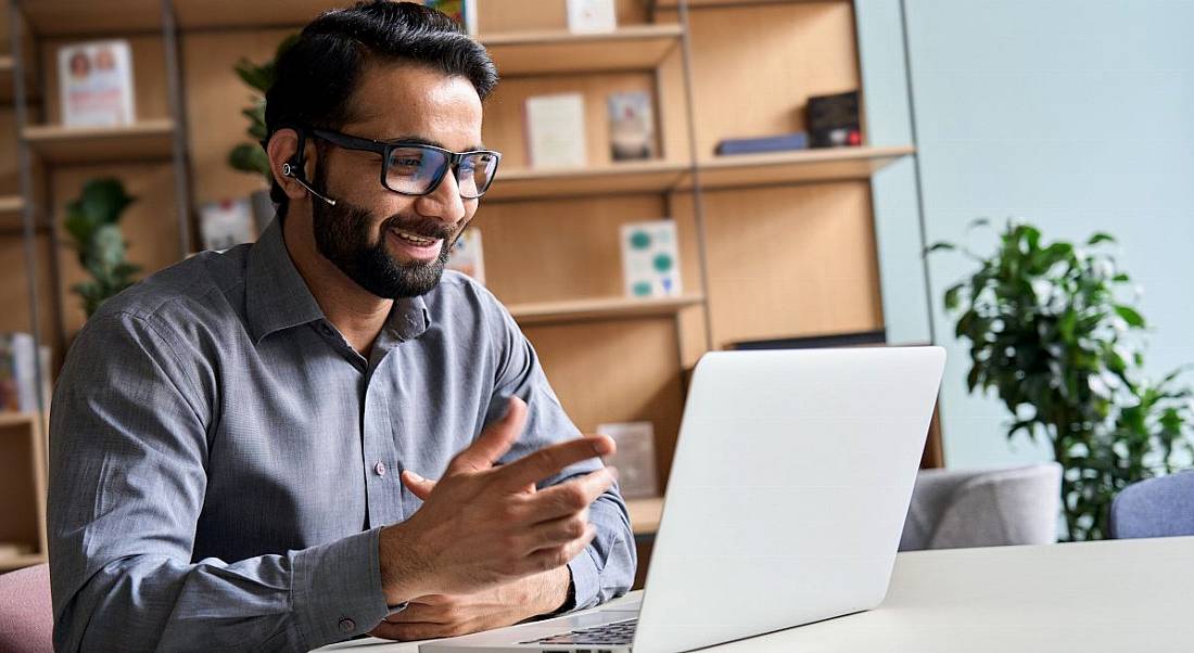 A man smiling working from home, sitting at a desk working on a laptop.