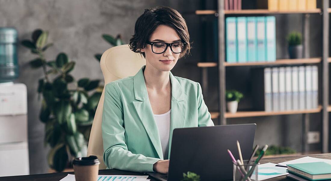 A woman in a mint green blazer sits at a desk in a brightly lit office working on a computer.