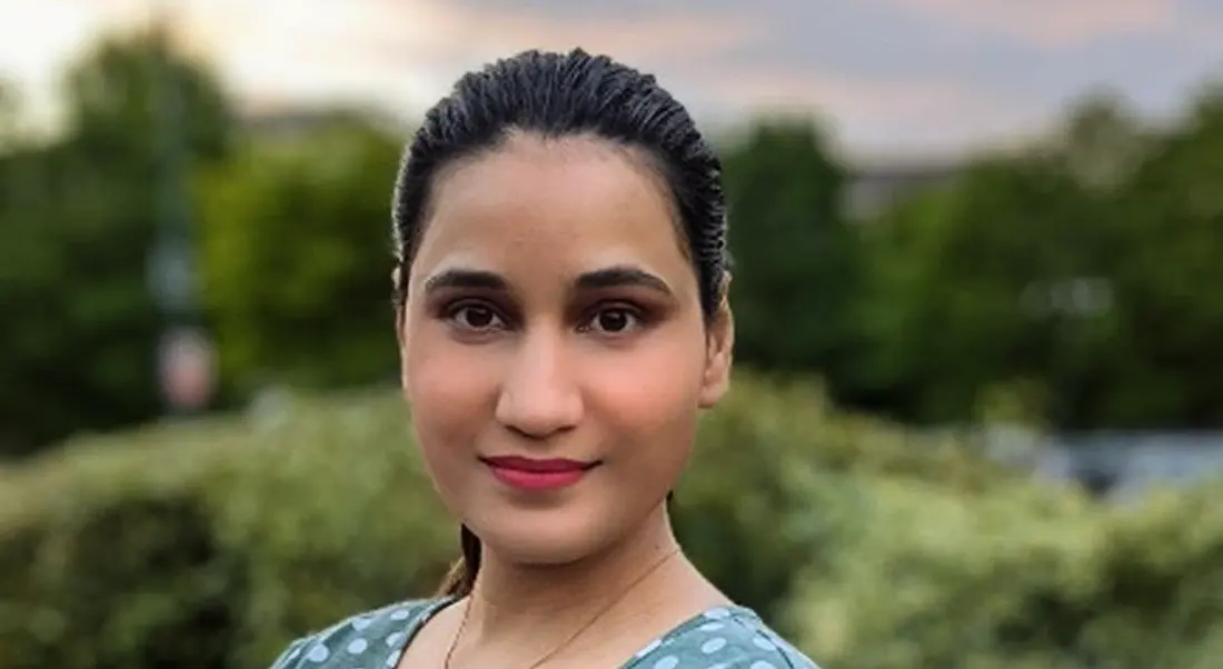 A close-up headshot of a woman smiling at the camera against a picturesque outdoor background.