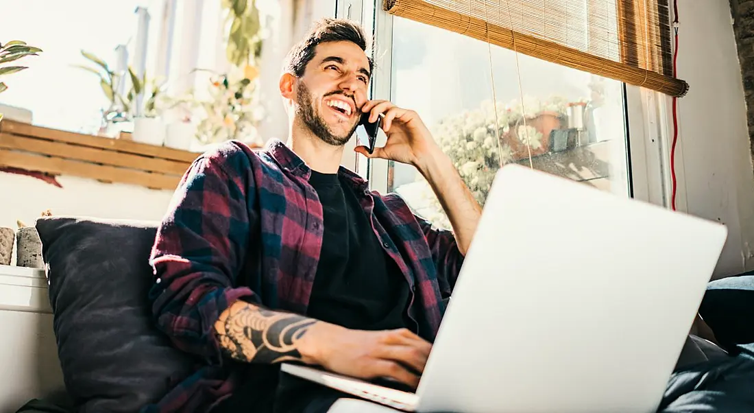 A man working from home taking a phone call while on a laptop. He’s seated upright on a bed and casually dressed.