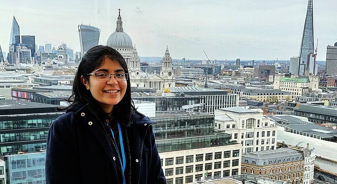 A woman stands against a wide city landscape with many tall buildings, smiling at the camera.