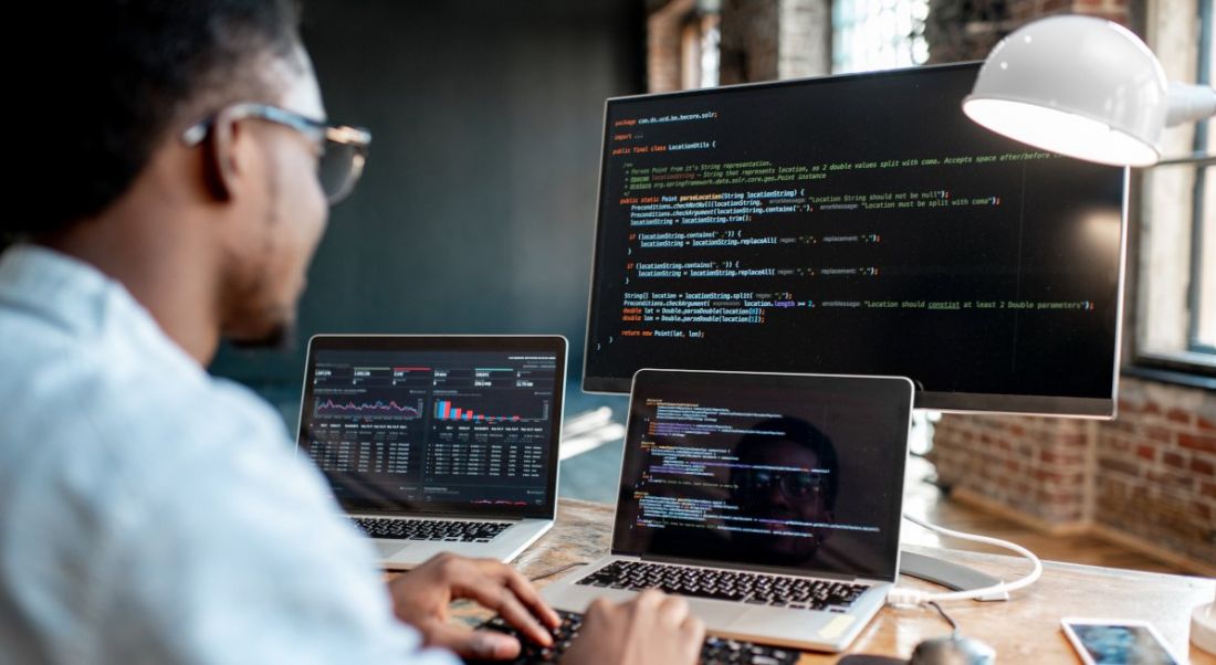 A software developer working at a large desk with two laptops and a large central screen, all displaying lines of code.