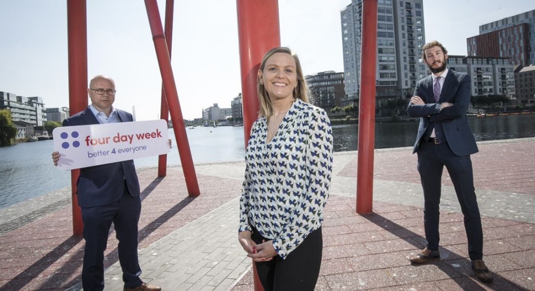 Three people stand beside a red sculpture in Dublin's Grand Canal Dock. One of the men is holding a sign that says 'Four Day Week'.