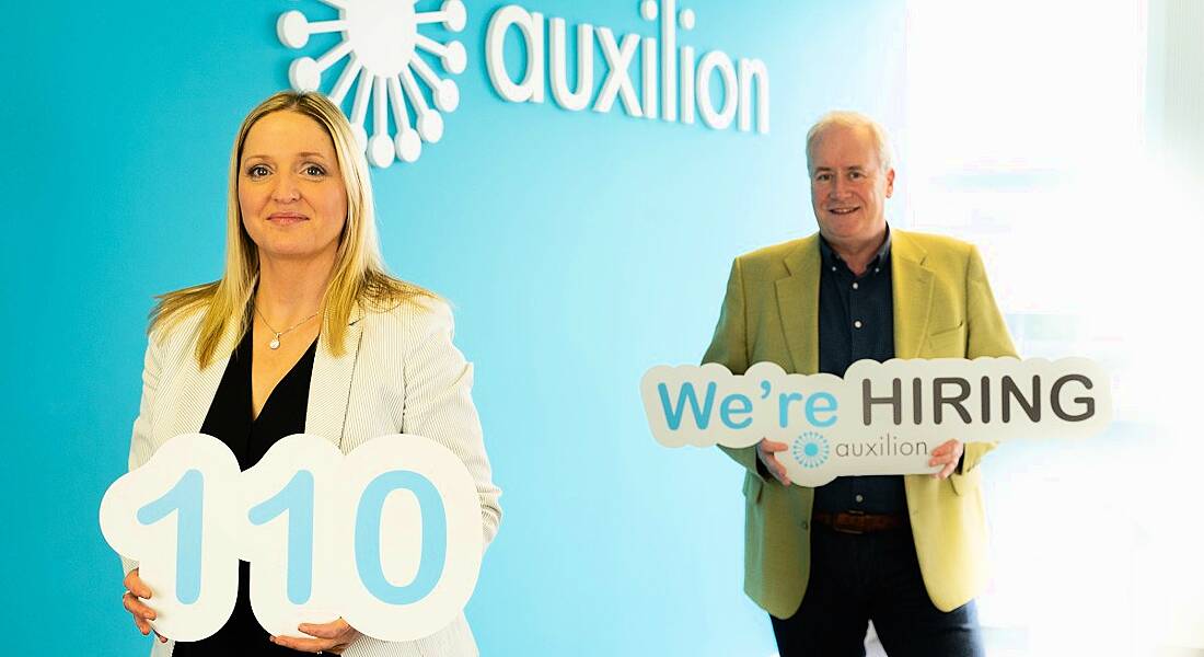 A woman and a man stand a few metres apart beside a blue wall with a white Auxilion logo on it. They’re holding signs - one that has the number 110 on it, and one that says 'We’re hiring'.