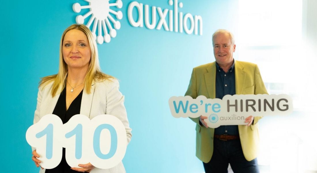 A woman and a man stand a few metres apart beside a blue wall with a white Auxilion logo on it. They’re holding signs - one that has the number 110 on it, and one that says 'We’re hiring'.