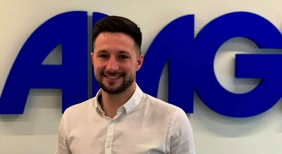 A young man in a white shirt stands in front of the blue Amgen logo on a white wall. He’s smiling at the camera.