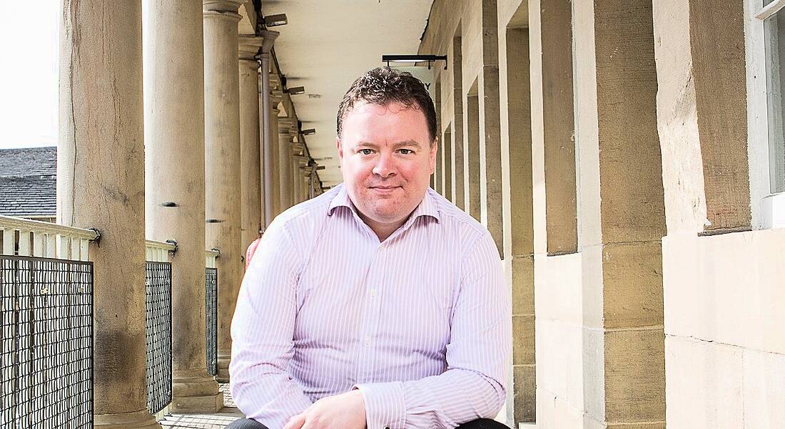 A man in a light coloured shirt faces the camera in an outdoor setting surrounded by stone pillars.