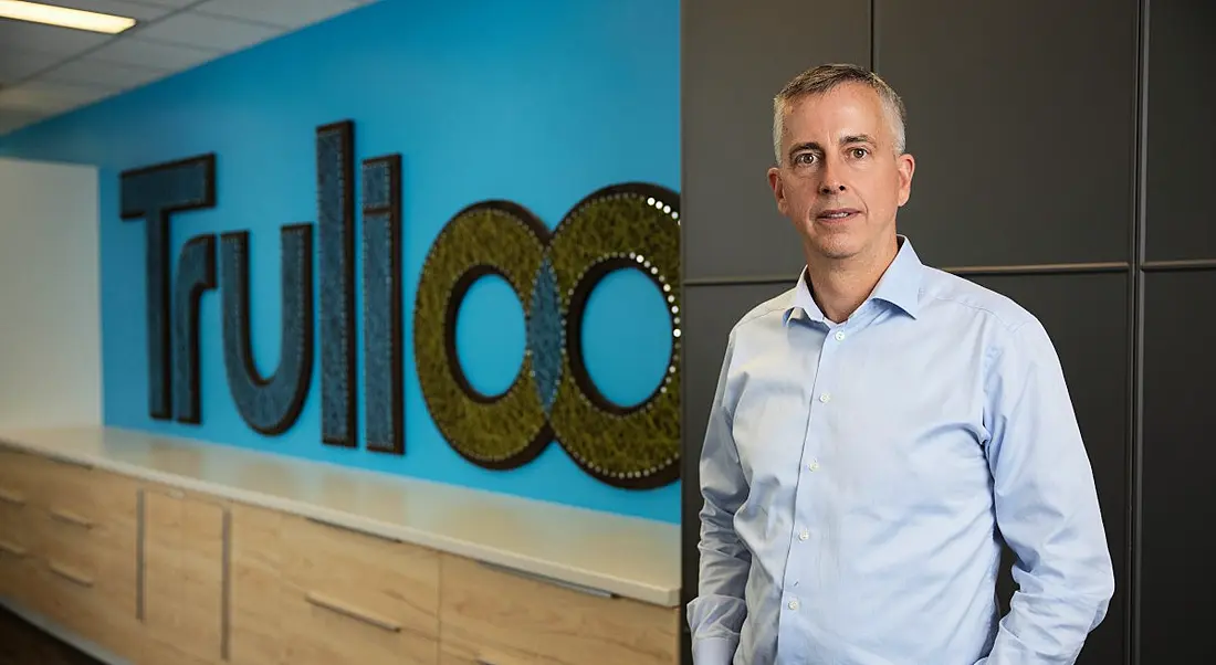 A man in a shirt stands in an office in front of a sign that says Trulioo.