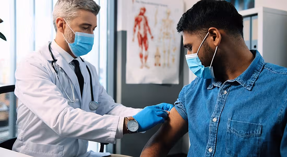 A man in a denim shirt wearing a face covering receives a vaccination from a doctor in a lab coat wearing a face covering.