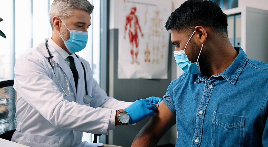 A man in a denim shirt wearing a face covering receives a vaccination from a doctor in a lab coat wearing a face covering.