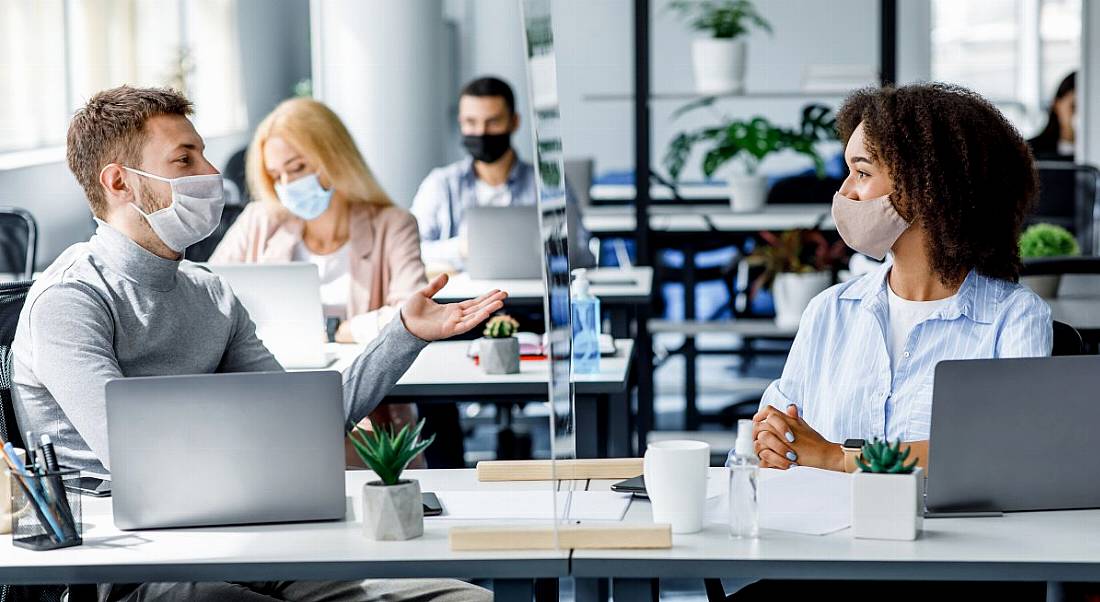 Four managers sit in an open-plan office wearing face coverings with protective screens between desks.