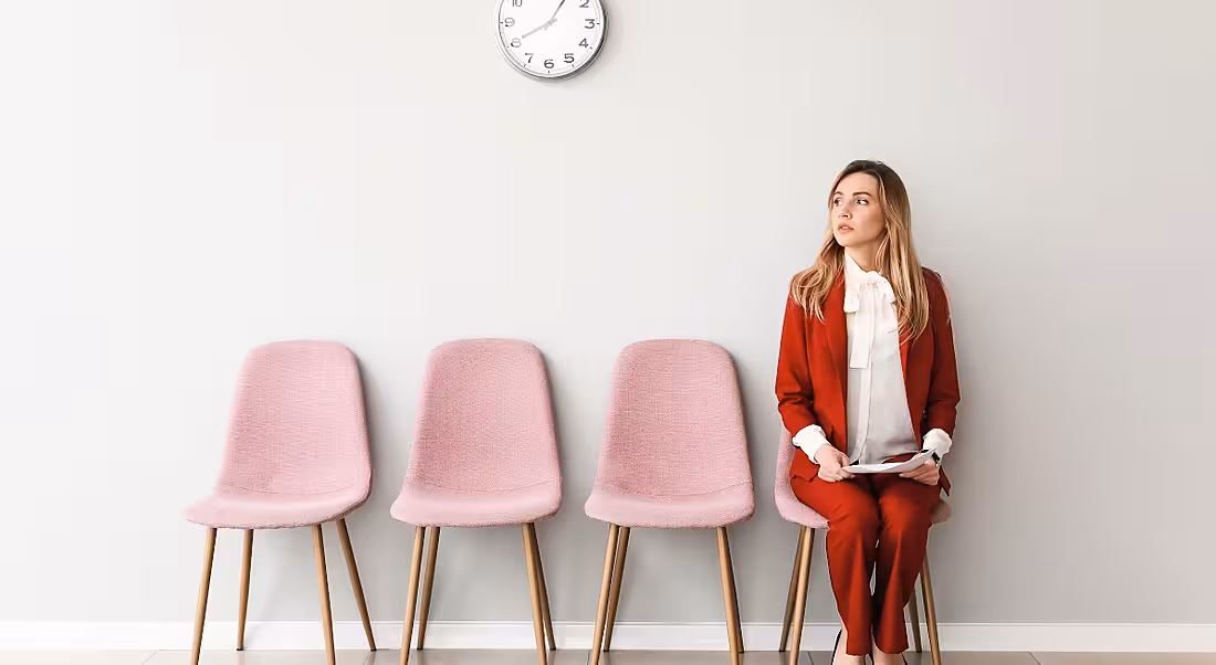 A woman in a red business suit sits at the end of a row of chairs nervously holding a CV before a job interview.