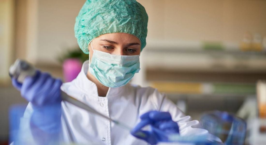 A woman working in a lab in a lab coat, mask and protective gear while using a pipette, highlighting pharma and healthcare careers.