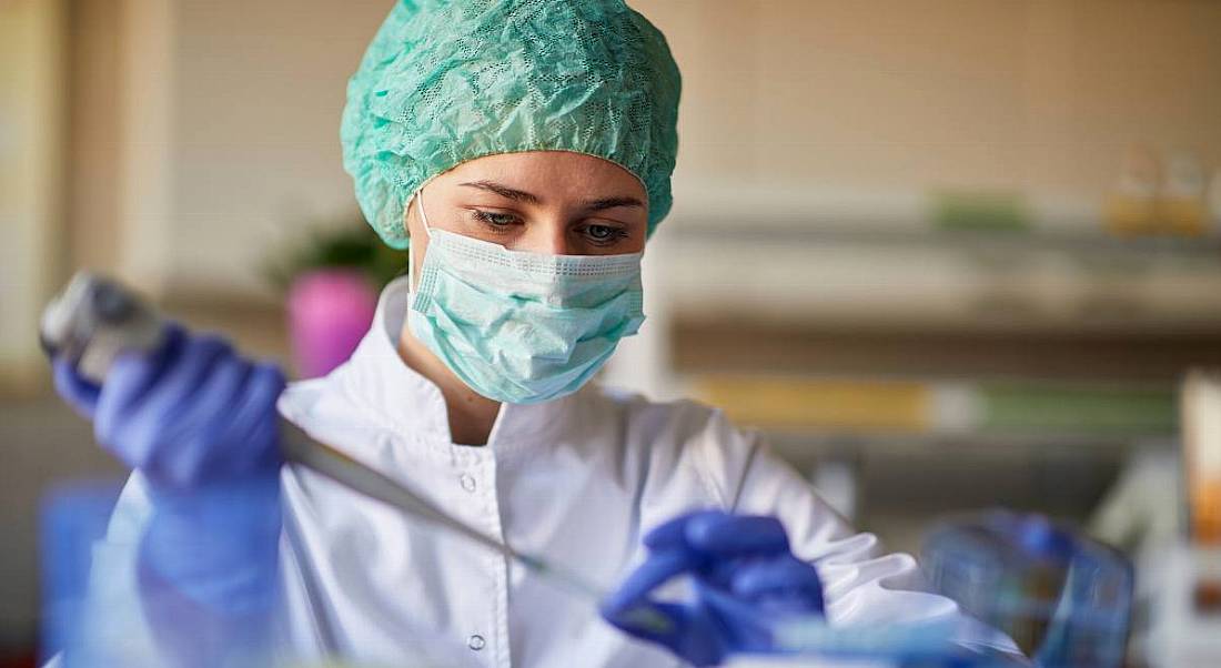 A woman working in a lab in a lab coat, mask and protective gear while using a pipette, highlighting pharma and healthcare careers.