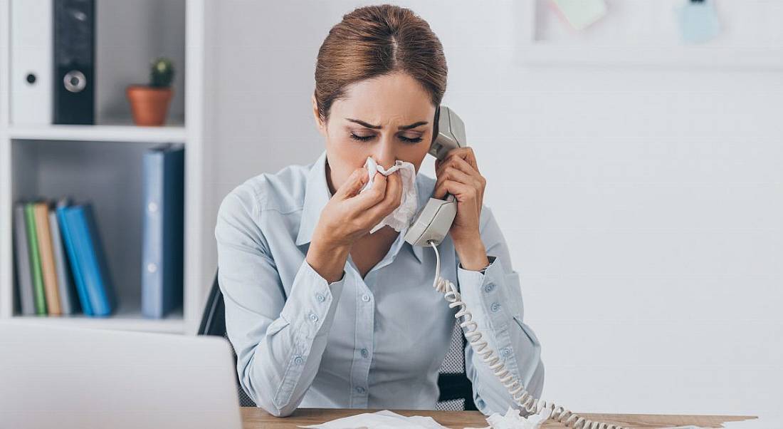 A woman sitting at her work desk using a phone while blowing her nose. Her desk is covered in used tissues.