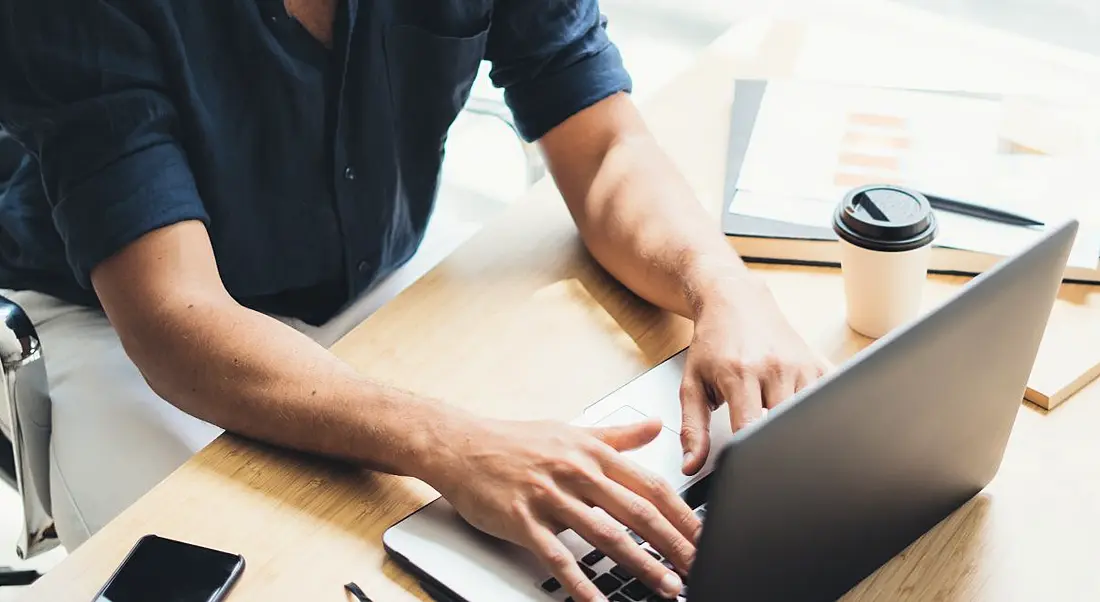 A close-up of a man’s hands working on a laptop in a brightly lit room with a coffee cup on the desk beside him.