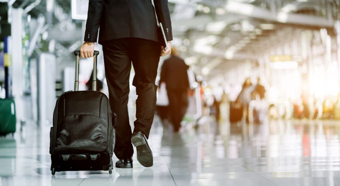 A man in a business suit walking through an airport wheeling a small suitcase behind him.