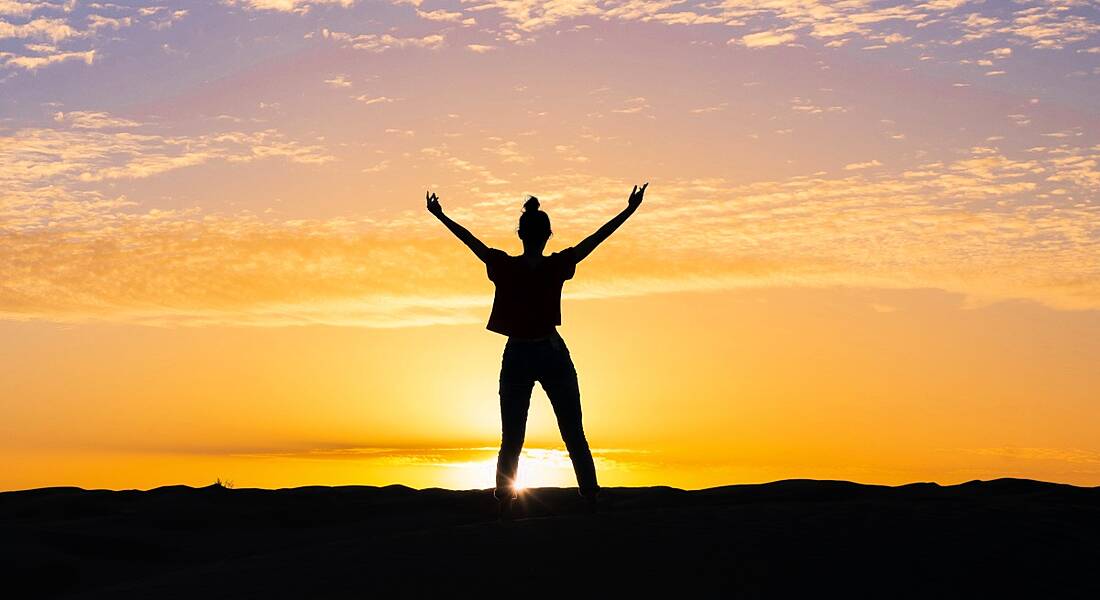 A silhouette of a woman standing with arms outstretched towards the sky at sunset.