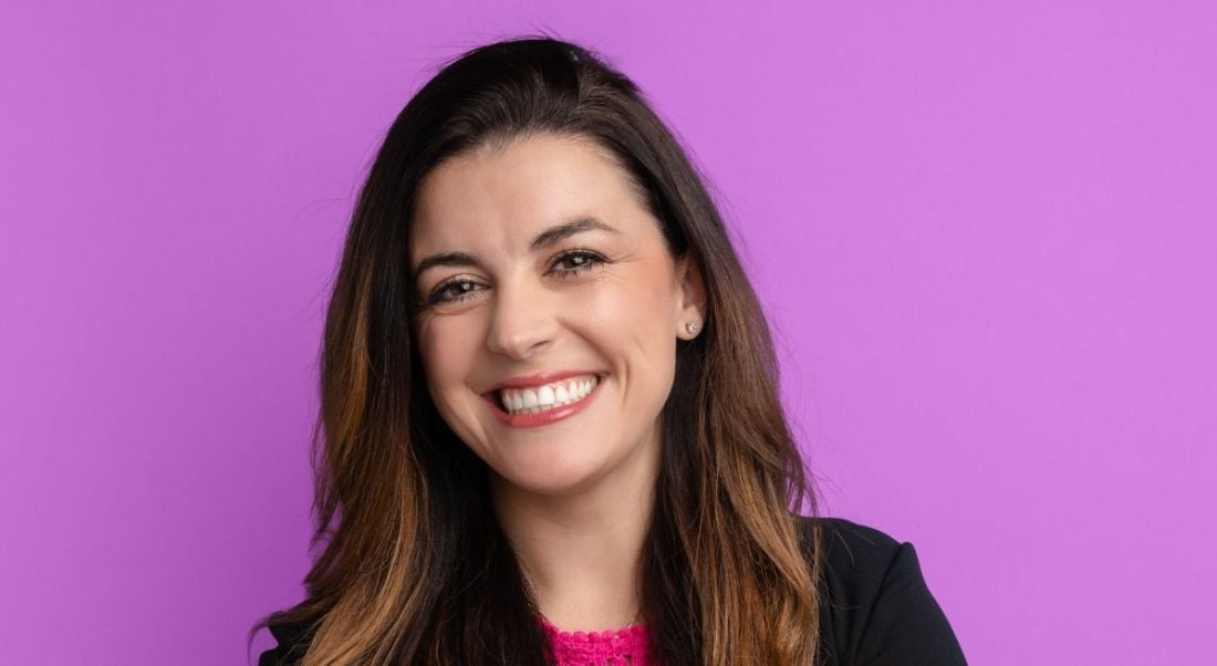 A close-up of Melanie Fellay, a woman with dark hair smiling at the camera against a bright purple background.