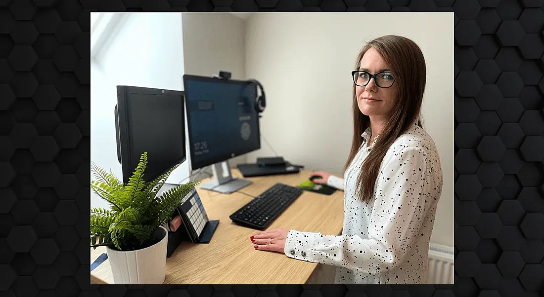 A woman with long brown hair and glasses stands at a desk with two monitors while looking at the camera.