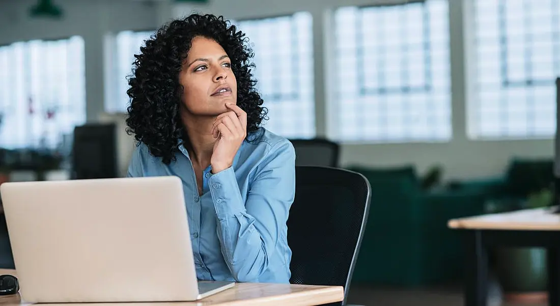 Young businesswoman looking deep in thought while sitting at her desk in a large modern office working online with a laptop.