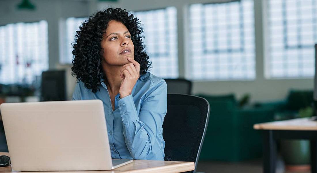 Young businesswoman looking deep in thought while sitting at her desk in a large modern office working online with a laptop.