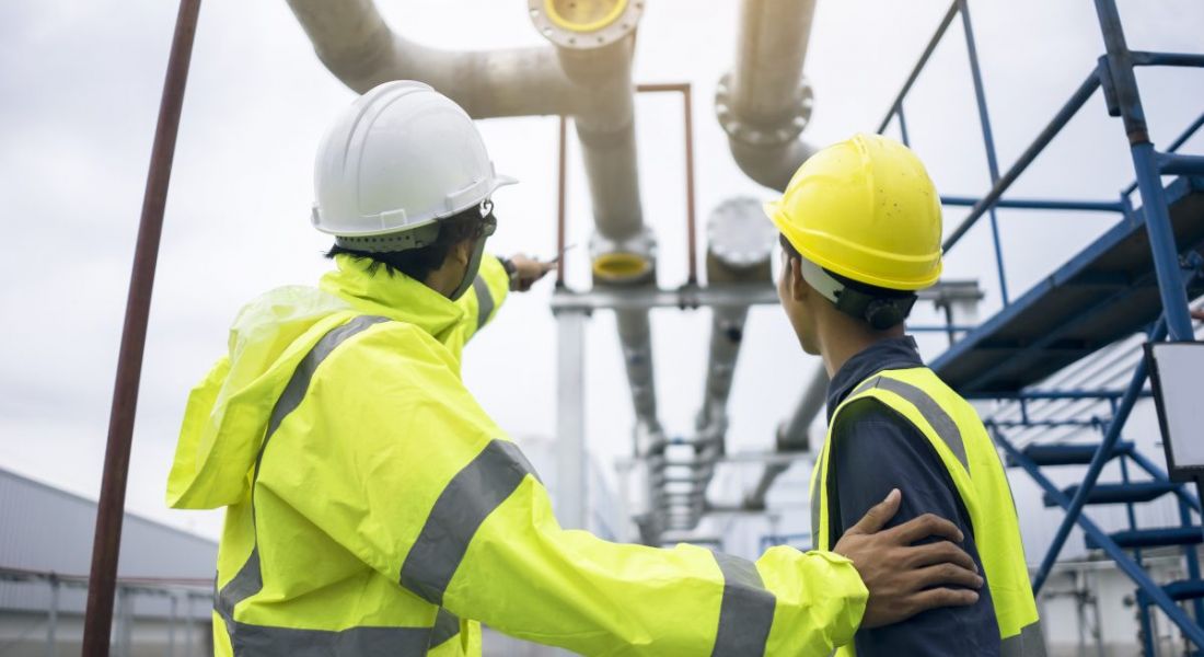 A senior engineer pats the arm of a junior colleague as they stand gazing up at a building site. Both are wearing protective fluorescent hardhats and jackets.