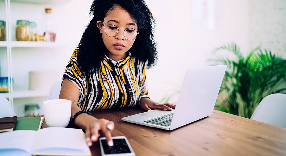 Young woman working at a desk with a laptop and smartphone and open notebook in front of her