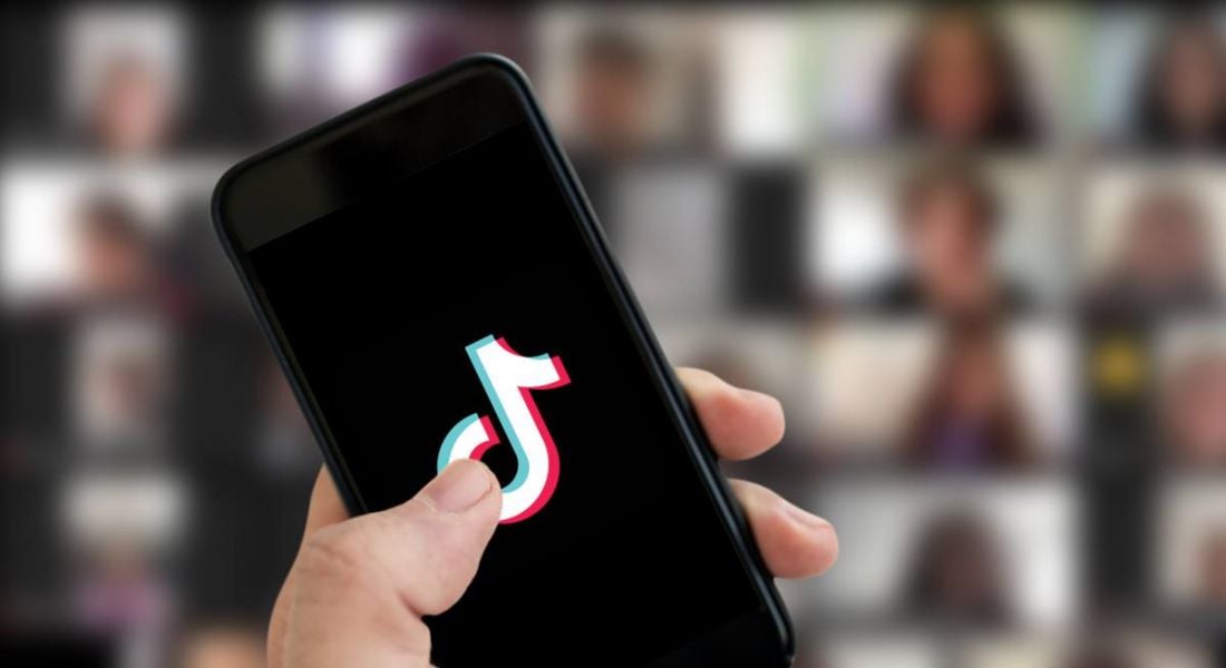 A hand holding up a smartphone with the TikTok logo on the screen. In the background, out of focus, is a larger screen displaying videos from a variety of people.