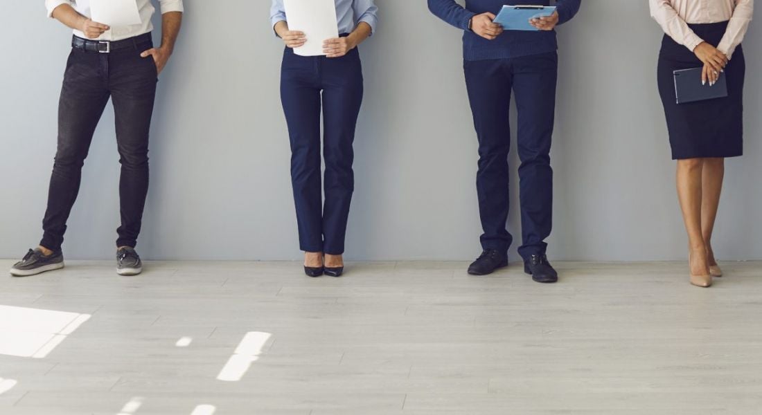 Four people waiting for a job interview. They are pictured from the waist down standing in a line in an empty neutral-coloured room.