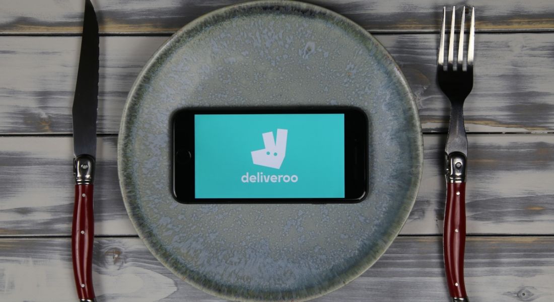 A dinner plate on a grey wooden table with a knife and fork either side. The dinner plate is empty except for a smartphone with the Deliveroo company graphic on its screen.
