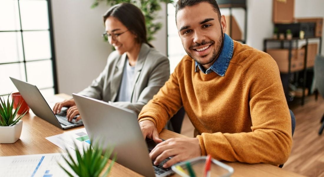 Two office workers sitting in an open-plan office working on their laptops, smiling.