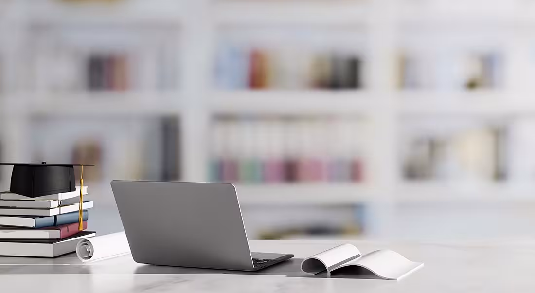 Pile of books with a graduation cap on top beside a laptop and an open book at a desk in a library or academic setting.
