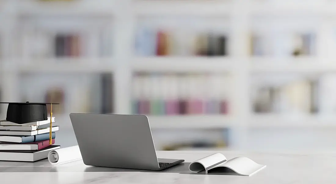 Pile of books with a graduation cap on top beside a laptop and an open book at a desk in a library or academic setting.