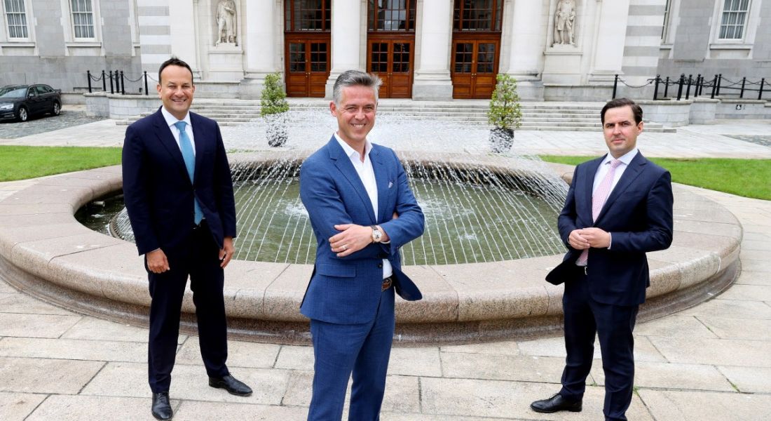Three men in suits stand, safely socially distanced, in front of a fountain outside Irish Government buildings.