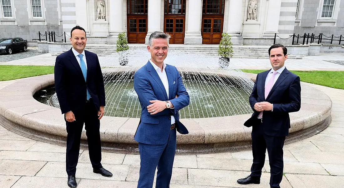 Three men in suits stand, safely socially distanced, in front of a fountain outside Irish Government buildings.