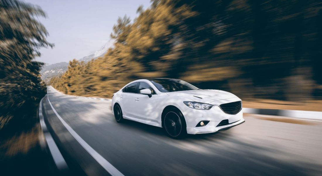 A white car speeds down a long, straight road surrounded by forestry on a sunny day.