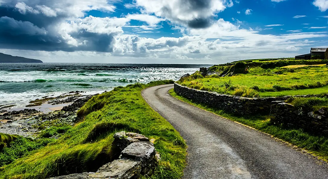 A winding Irish country road is shown. There is a small house on the right and the ocean is on the left of the road. There is a stone wall running alongside the road and there are fields of grass beyond the wall.