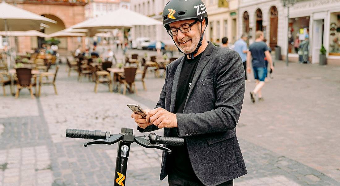 A man wearing a helmet stands on a city street using his phone, with an e-scooter beside him.