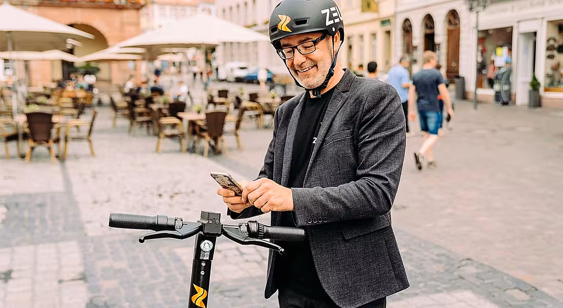 A man wearing a helmet stands on a city street using his phone, with an e-scooter beside him.
