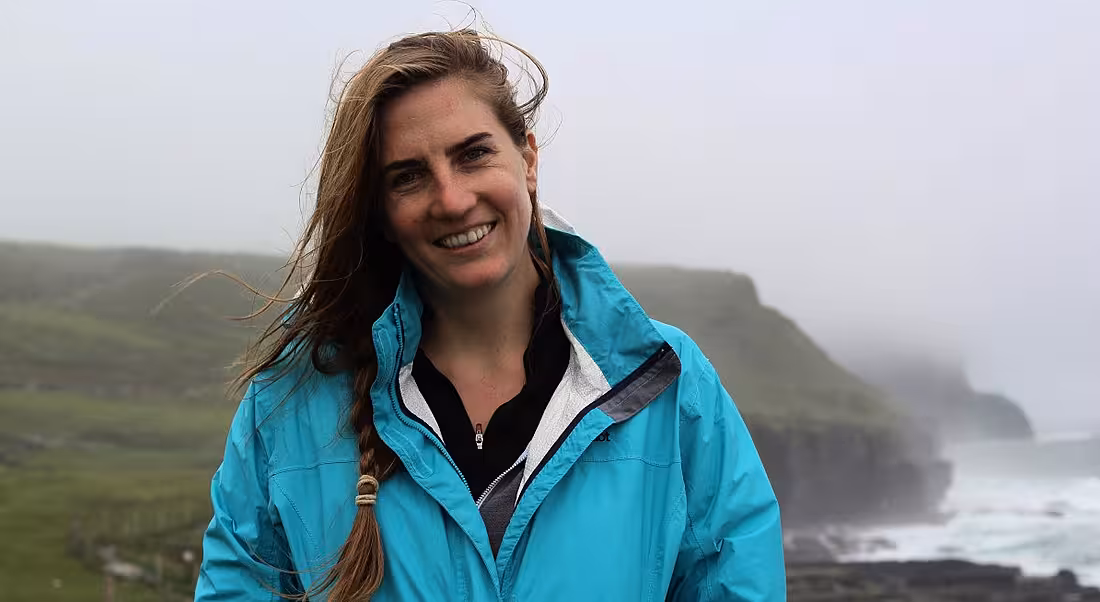 A young woman in a bright blue windbreaker stands on a hilltop on a windy, foggy day, smiling at the camera.
