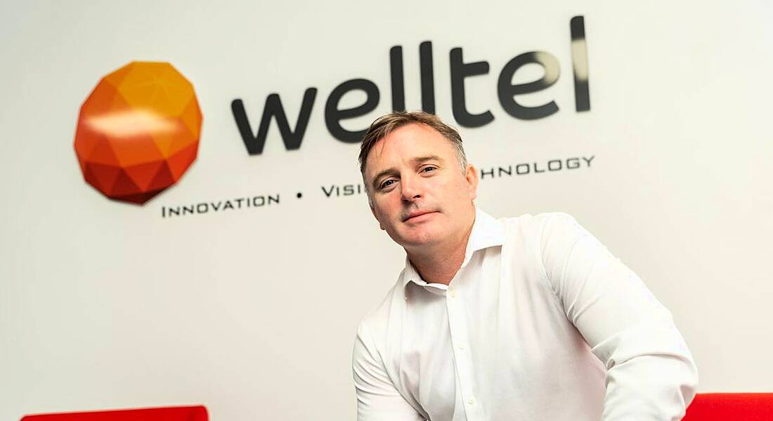 A man in a white shirt sits on a red chair. On the wall behind him is the Welltel logo.